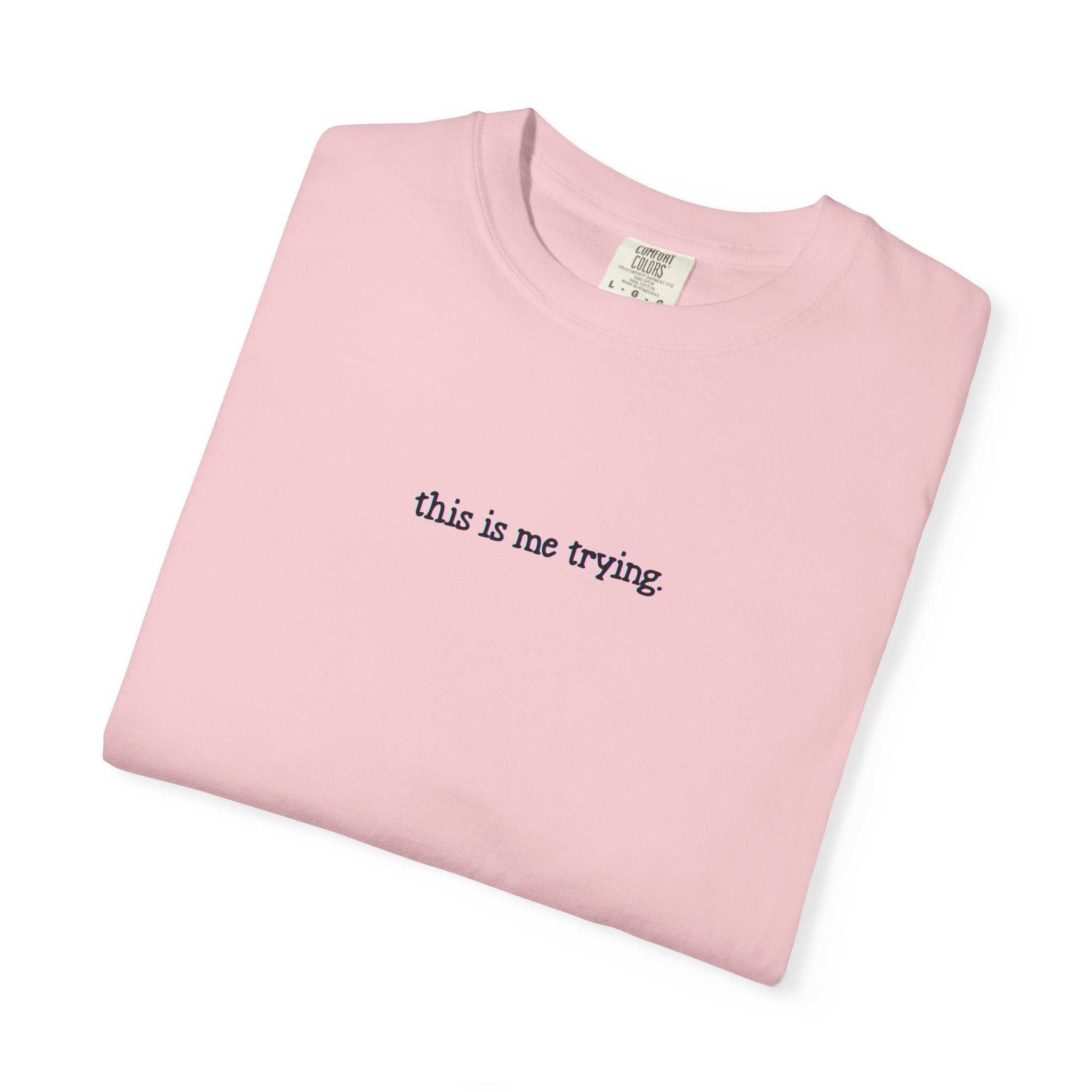 Folded pink t-shirt with 'this is me trying' text on a white background
