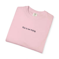 Folded pink t-shirt with 'this is me trying' text on a white background