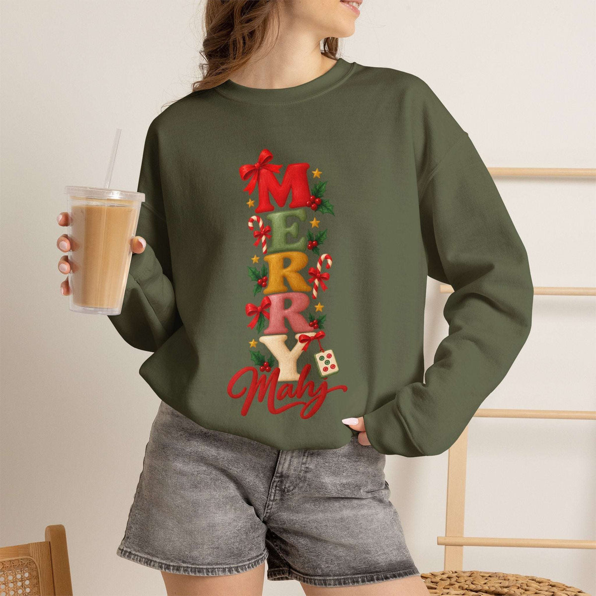 Female model with ice coffee wearing a Military green Merry Mahj Christmas mahjong sweatshirt with colorful stacked lettering, red bows, candy canes, holly and a mahjong tile.