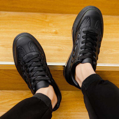 Black trendy leather men's casual sneakers with car stitching and rubber sole, worn with black pants on wooden stairs