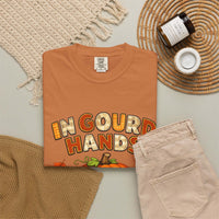 A folded terracotta orange t-shirt with a 'In Gourd Hands' Thanksgiving graphic, styled in a flat lay with beige pants, a knit blanket, a jar candle, and a woven placemat on a light gray surface.