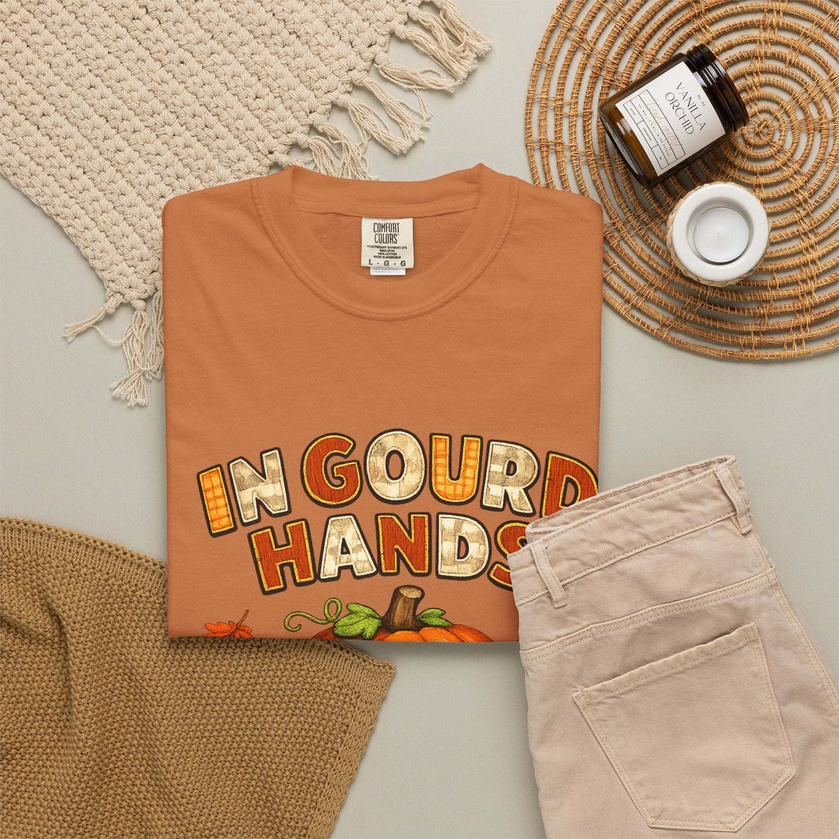 A folded terracotta orange t-shirt with a 'In Gourd Hands' Thanksgiving graphic, styled in a flat lay with beige pants, a knit blanket, a jar candle, and a woven placemat on a light gray surface.