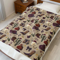 Plush blanket with cowboy Christmas design featuring boots, hats, cacti, and holiday elements on bed
