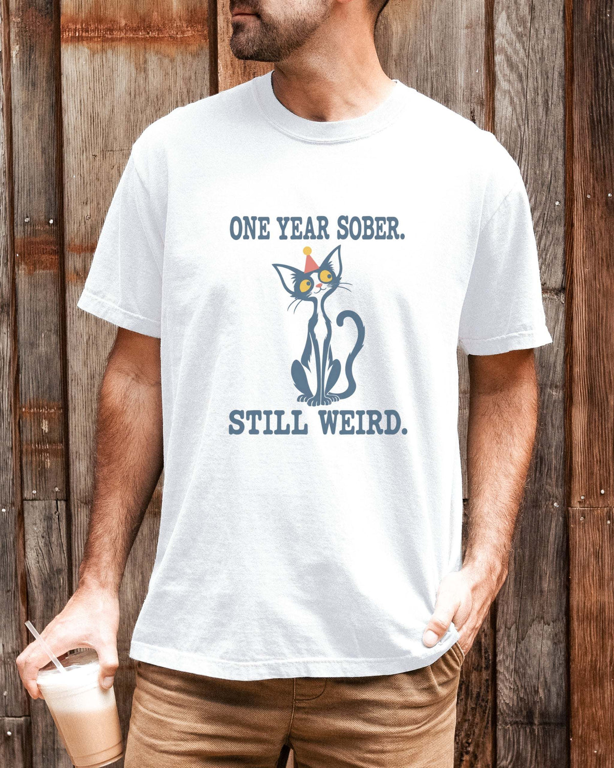 Male model wearing a white Comfort Colors 1717 t-shirt featuring a navy cat in a red party hat and “One Year Sober. Still Weird.” lettering.