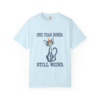Flat lay of a chambray blue Comfort Colors 1717 t-shirt featuring a navy cat in a red party hat and “One Year Sober. Still Weird.” lettering.
