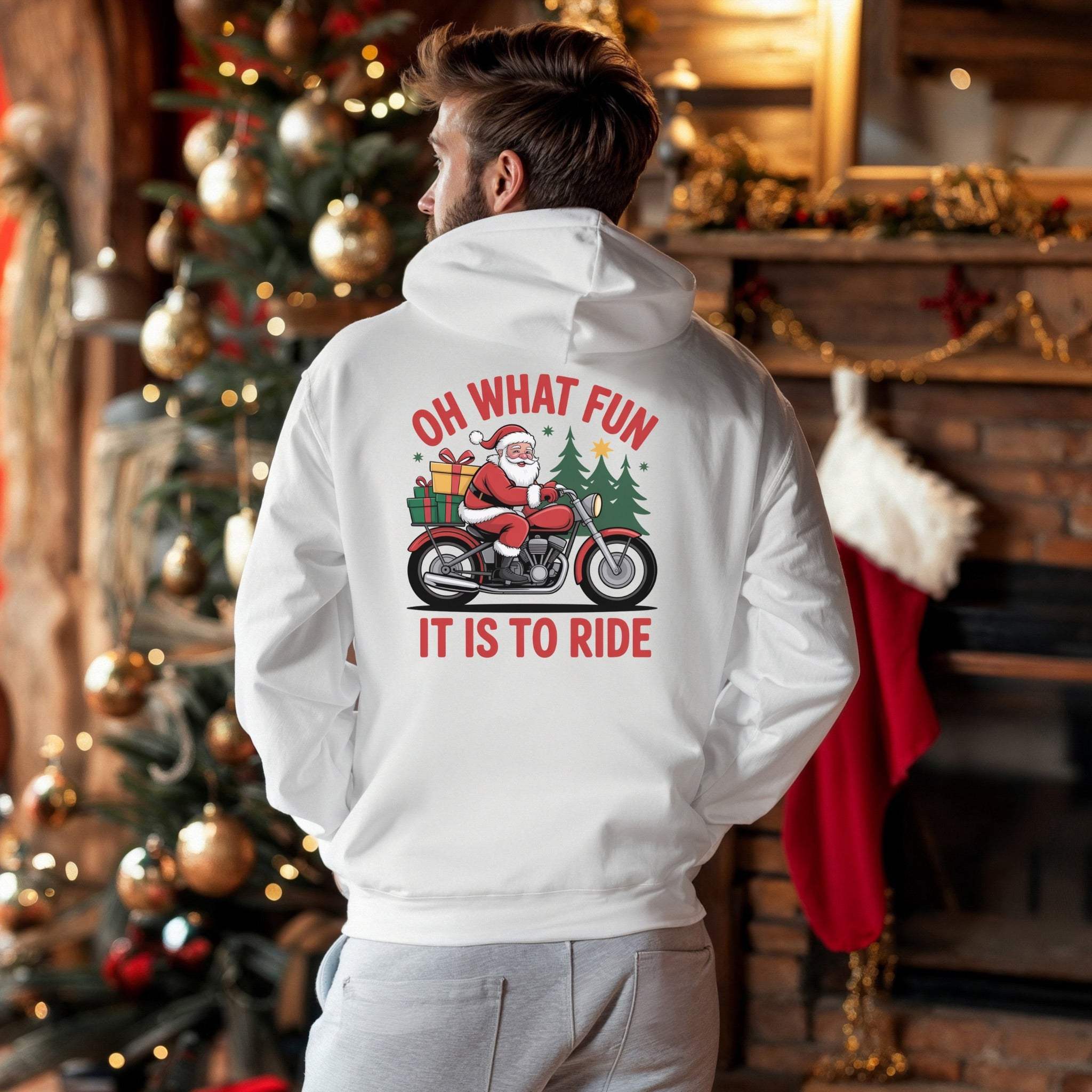 White Santa Motorcycle Hoodie with festive Santa on bike graphic and text "Oh What Fun It Is To Ride" worn indoors by man near Christmas tree and fireplace