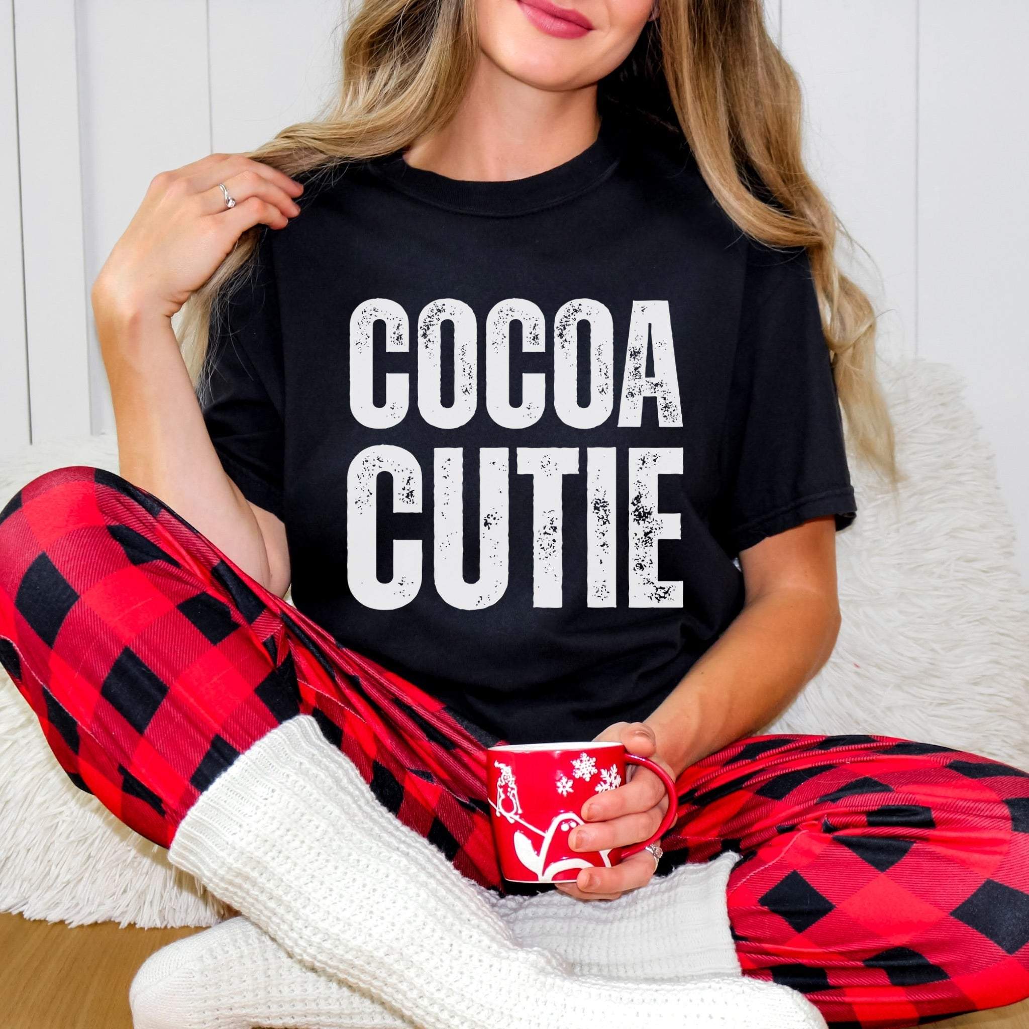 Woman wearing black Distressed Cocoa Cutie Tee with white text and red plaid pants holding a holiday mug