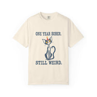 Flat lay of an ivory Comfort Colors 1717 t-shirt featuring a navy cat in a red party hat and “One Year Sober. Still Weird.” lettering.