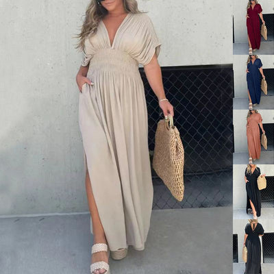 Woman wearing a beige bat-sleeved V-neck dress with elastic waist and side slit, holding a woven bag, standing against a light wall.