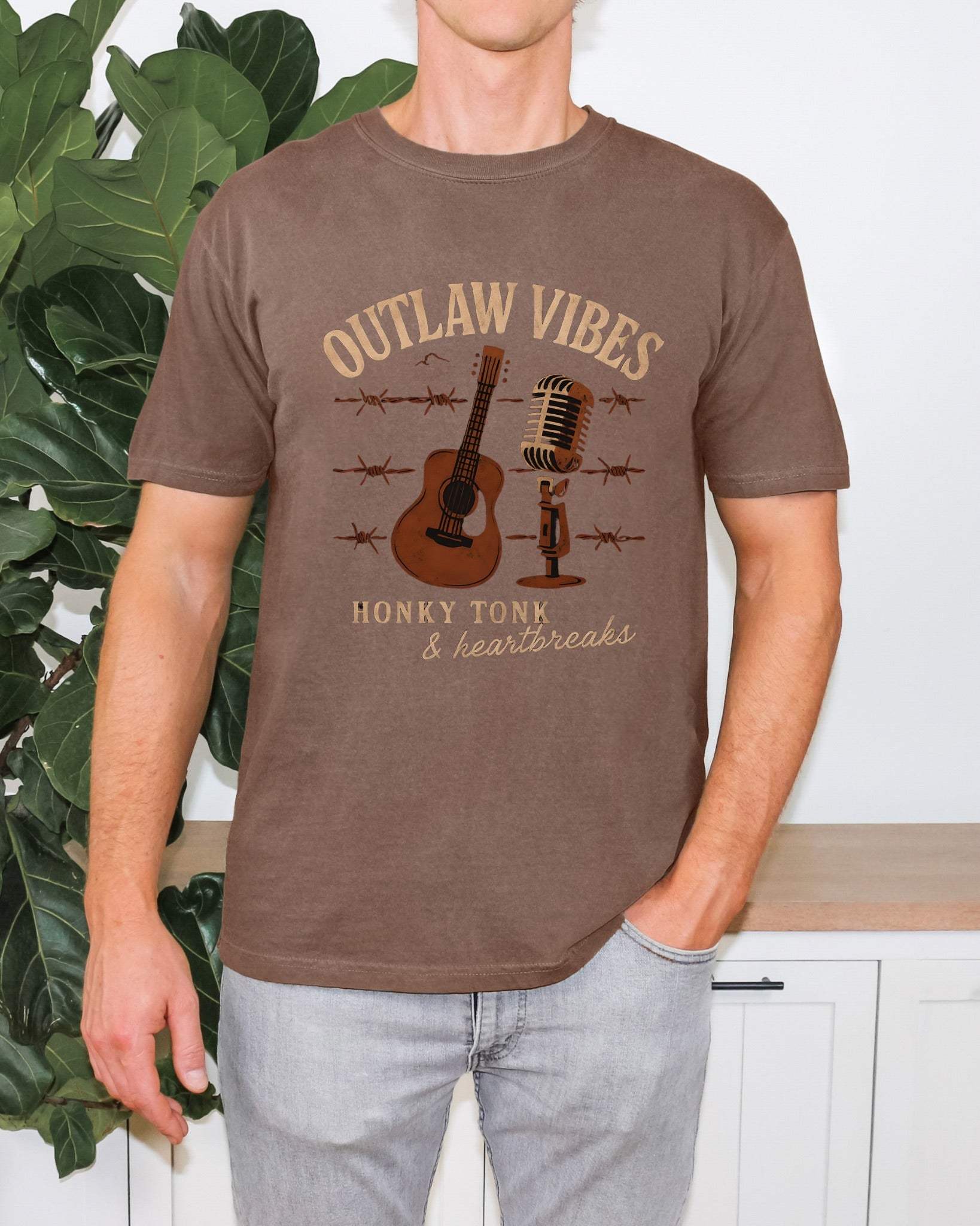 Man wearing brown Outlaw Vibes T-Shirt with guitar and microphone graphic, paired with light gray jeans