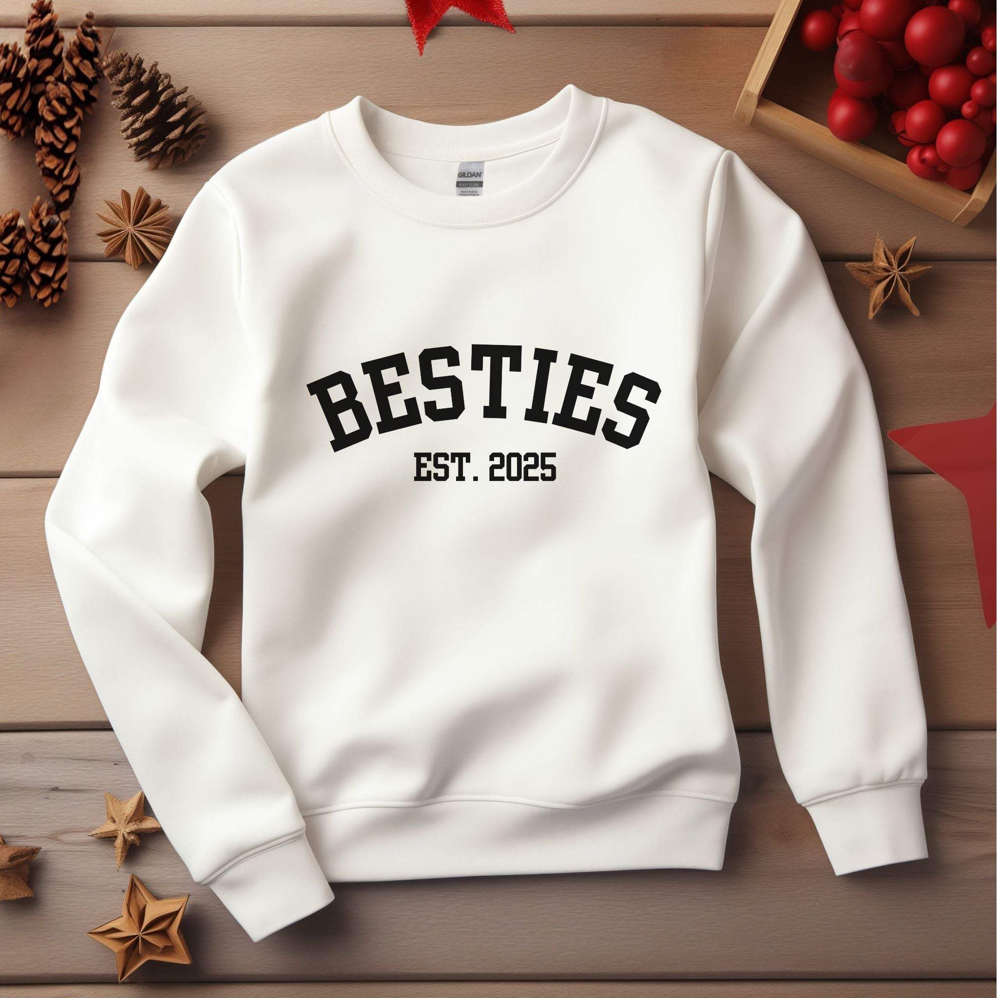 White Varsity Besties Crewneck Sweatshirt with black "BESTIES EST. 2025" lettering on a wooden background