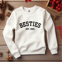 White Varsity Besties Crewneck Sweatshirt with black "BESTIES EST. 2025" lettering on a wooden background