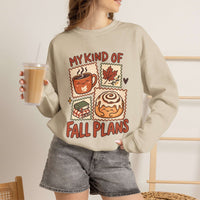 A woman wearing a beige crewneck sweatshirt with a colorful graphic that reads 'MY KIND OF FALL PLANS' above and below illustrations of a coffee mug, maple leaf, gift box, and cinnamon roll. She is holding an iced coffee and wearing denim shorts in a brightly lit indoor setting.