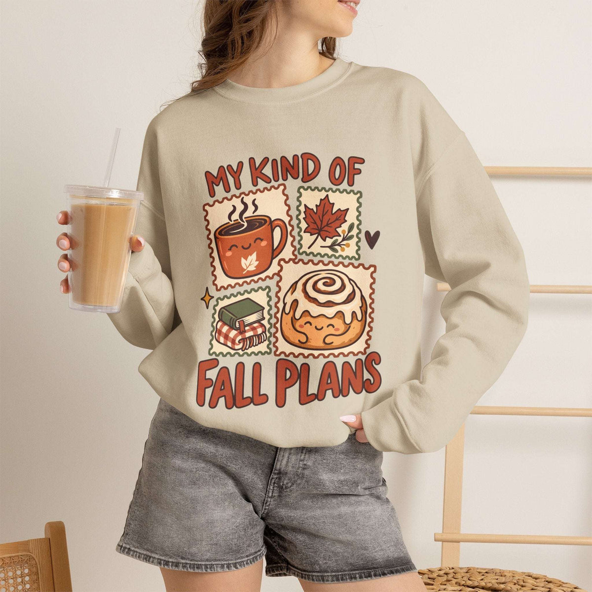 A woman wearing a beige crewneck sweatshirt with a colorful graphic that reads 'MY KIND OF FALL PLANS' above and below illustrations of a coffee mug, maple leaf, gift box, and cinnamon roll. She is holding an iced coffee and wearing denim shorts in a brightly lit indoor setting.