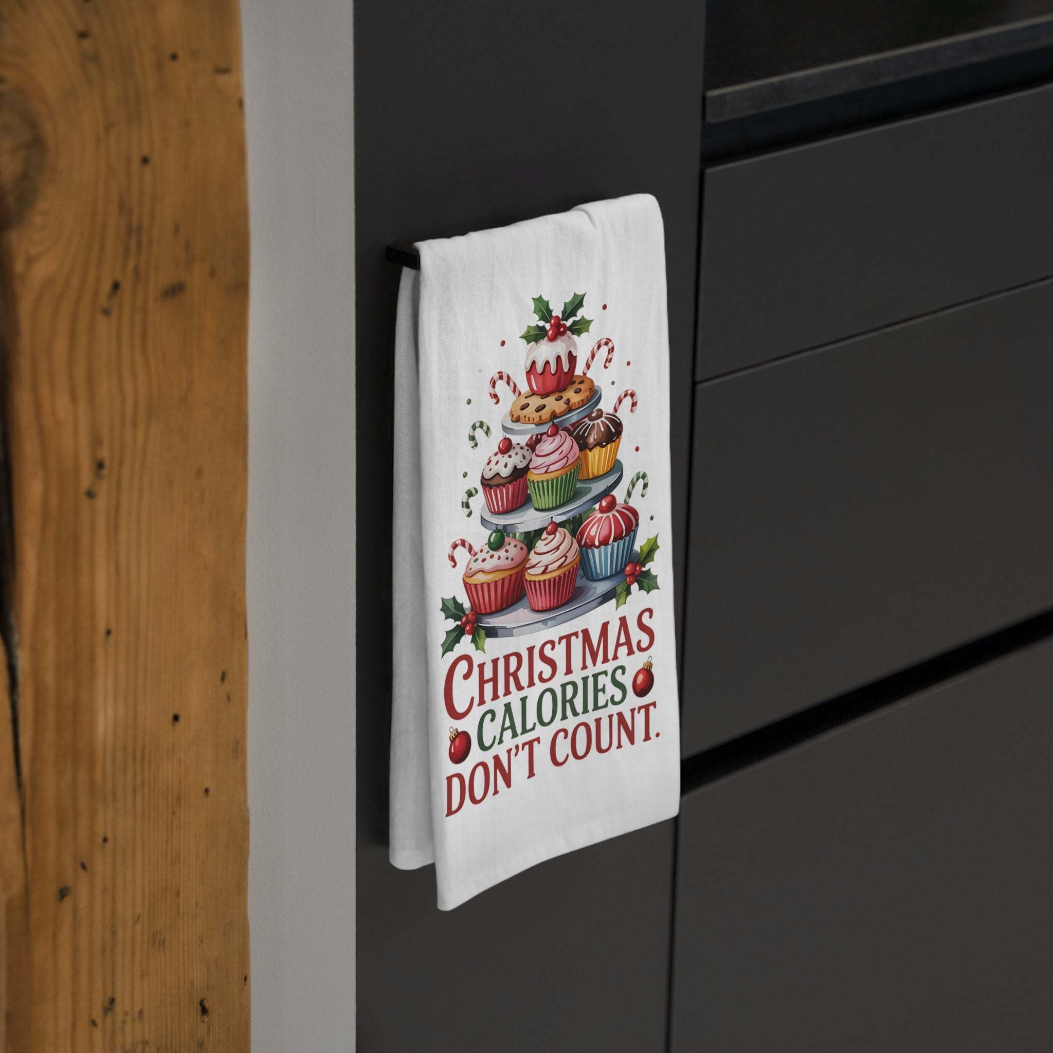White kitchen towel Christmas with festive cupcakes and "Christmas Calories Don't Count" text hanging on a black cabinet