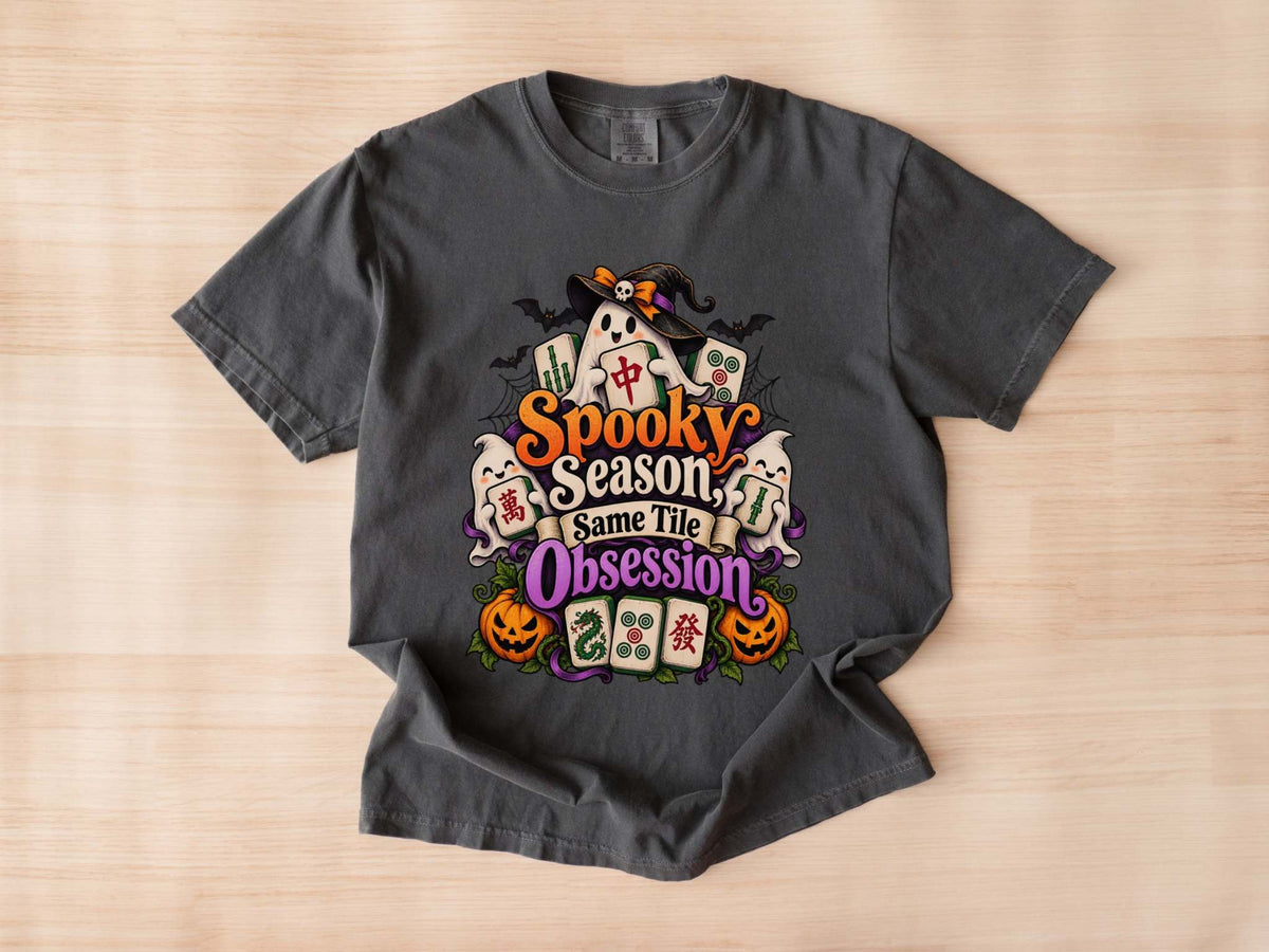 Flat lay of a pepper Halloween mahjong T-shirt with ghosts, pumpkins, mahjong tiles and “Spooky Season, Same Tile Obsession” lettering.