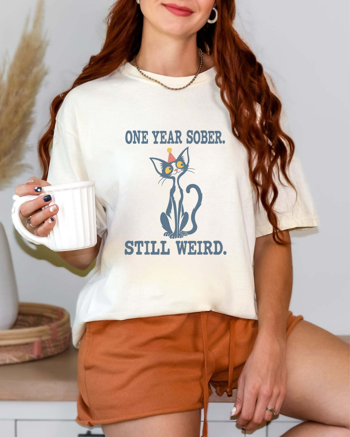 Female model wearing an Ivory Comfort Colors 1717 t-shirt featuring a navy cat in a red party hat and “One Year Sober. Still Weird.” lettering.