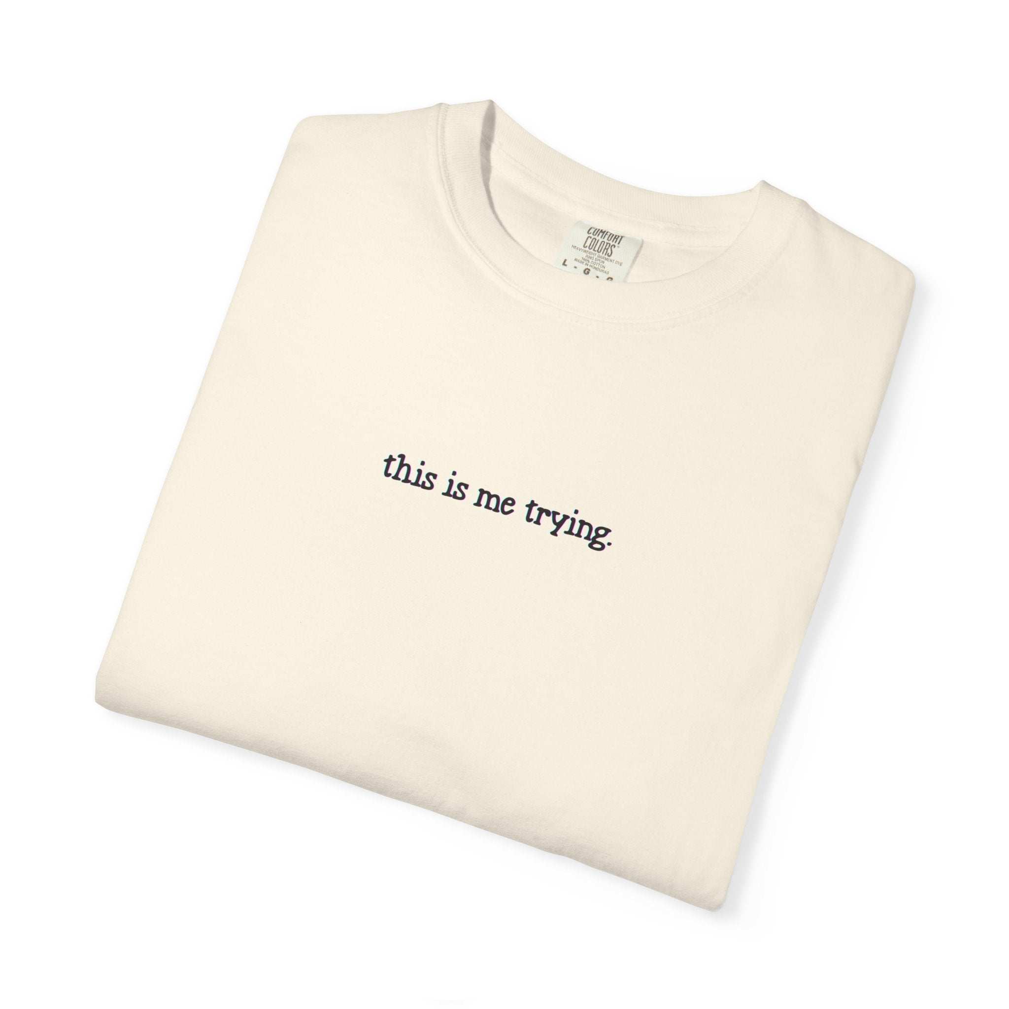 Folded beige t-shirt with 'this is me trying' text on a white background