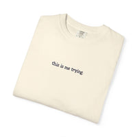 Folded beige t-shirt with 'this is me trying' text on a white background