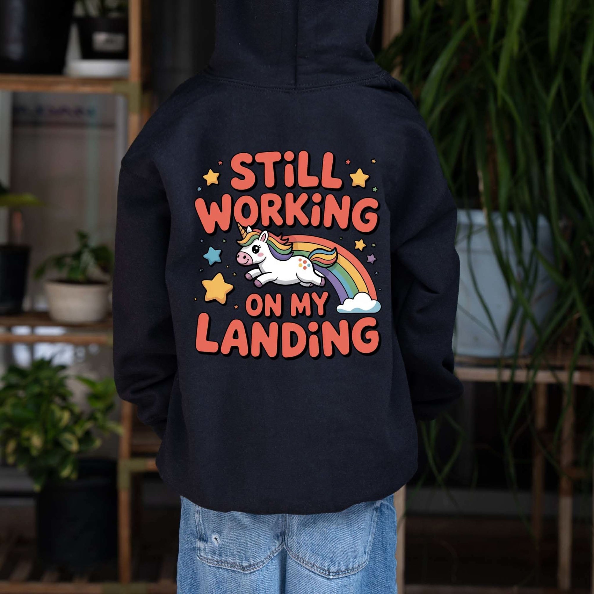 Kids hoodie with bright unicorn rainbow design and playful "Still Working On My Landing" text