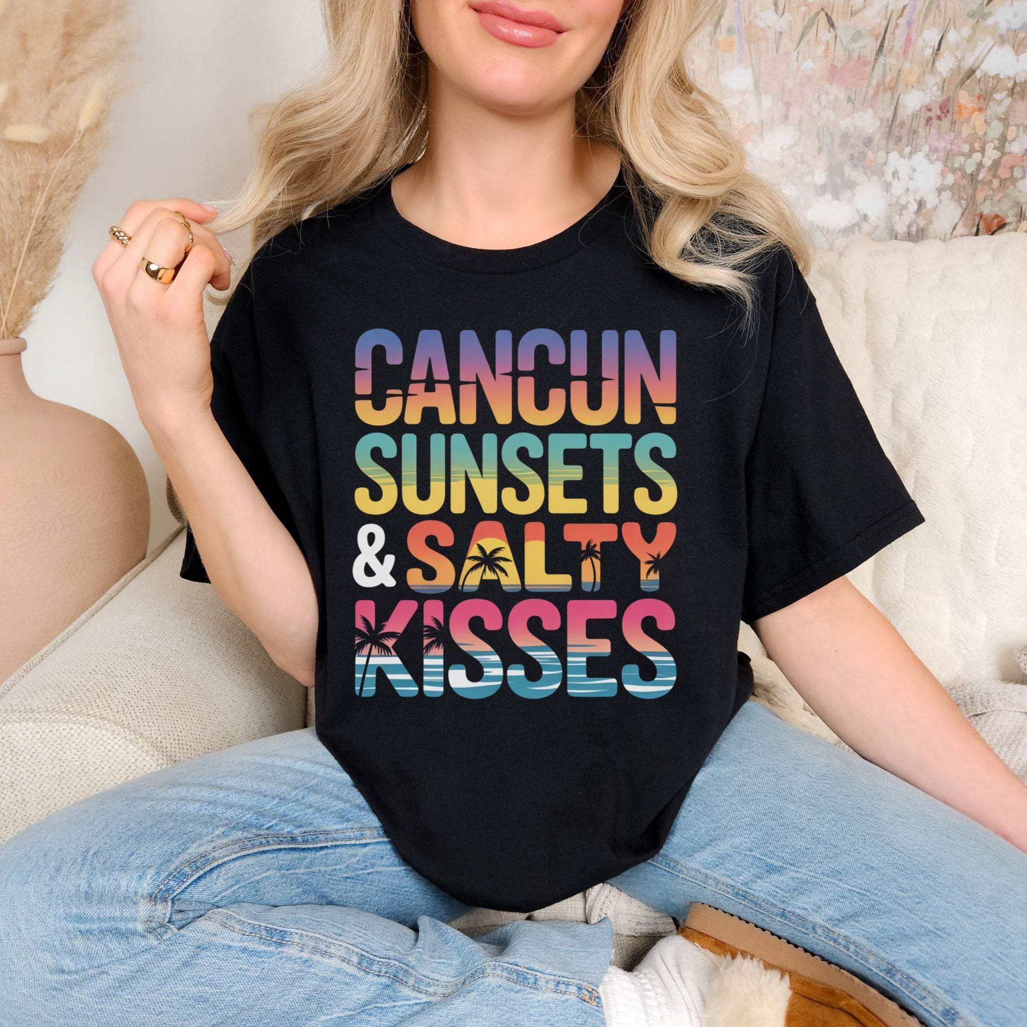 Black Cancun Sunsets and Salty Kisses tropical beach vacation t-shirt with colorful text worn by woman with blonde hair and jeans sitting indoors