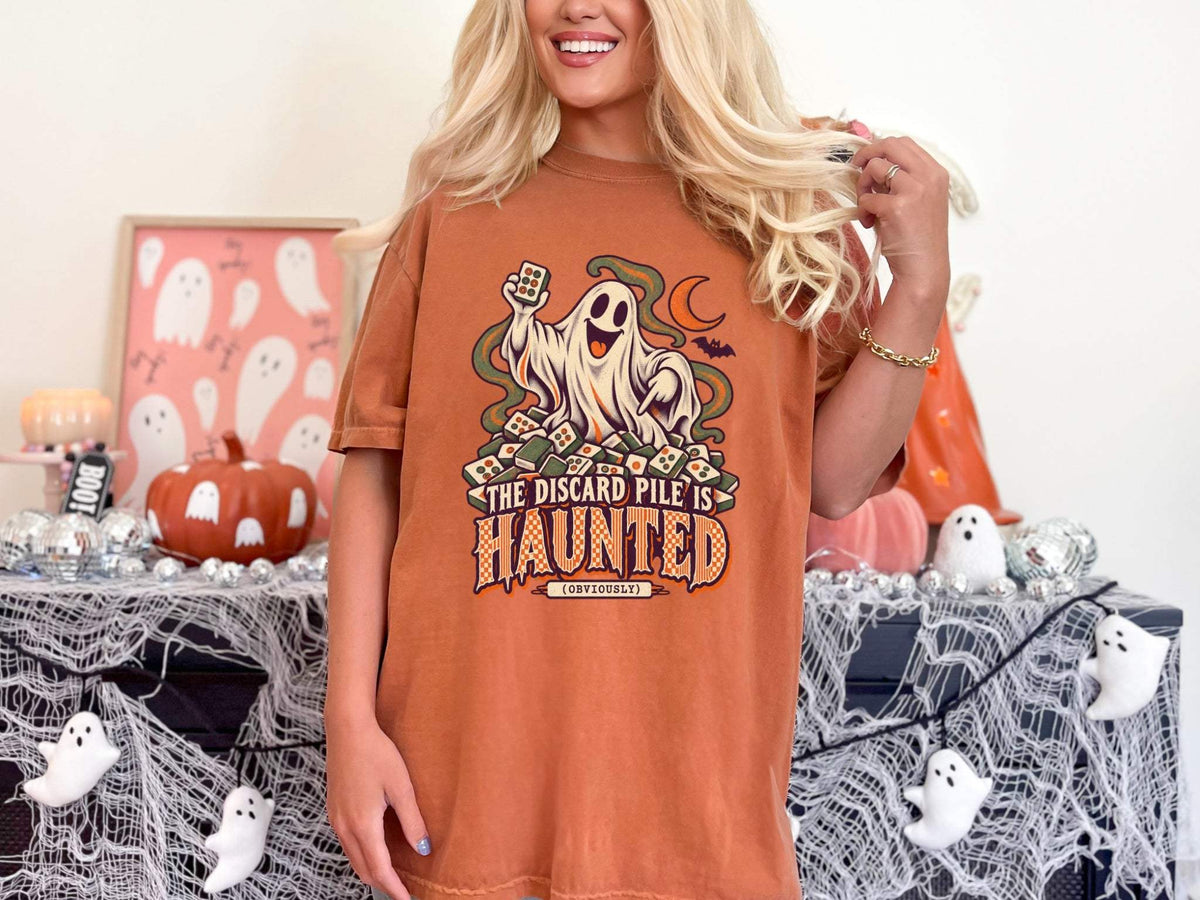 Model wearing yam orange Comfort Colors 1717 Halloween mahjong T-shirt with a ghost holding a tile and “The Discard Pile Is Haunted (Obviously)” lettering.