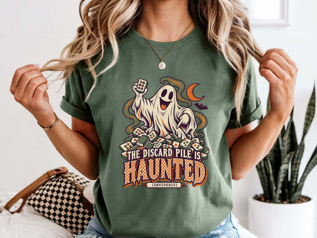 Model wearing moss green Comfort Colors 1717 Halloween mahjong T-shirt with a ghost holding a tile and “The Discard Pile Is Haunted (Obviously)” lettering.