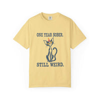 Flat lay of an butter Comfort Colors 1717 t-shirt featuring a navy cat in a red party hat and “One Year Sober. Still Weird.” lettering.