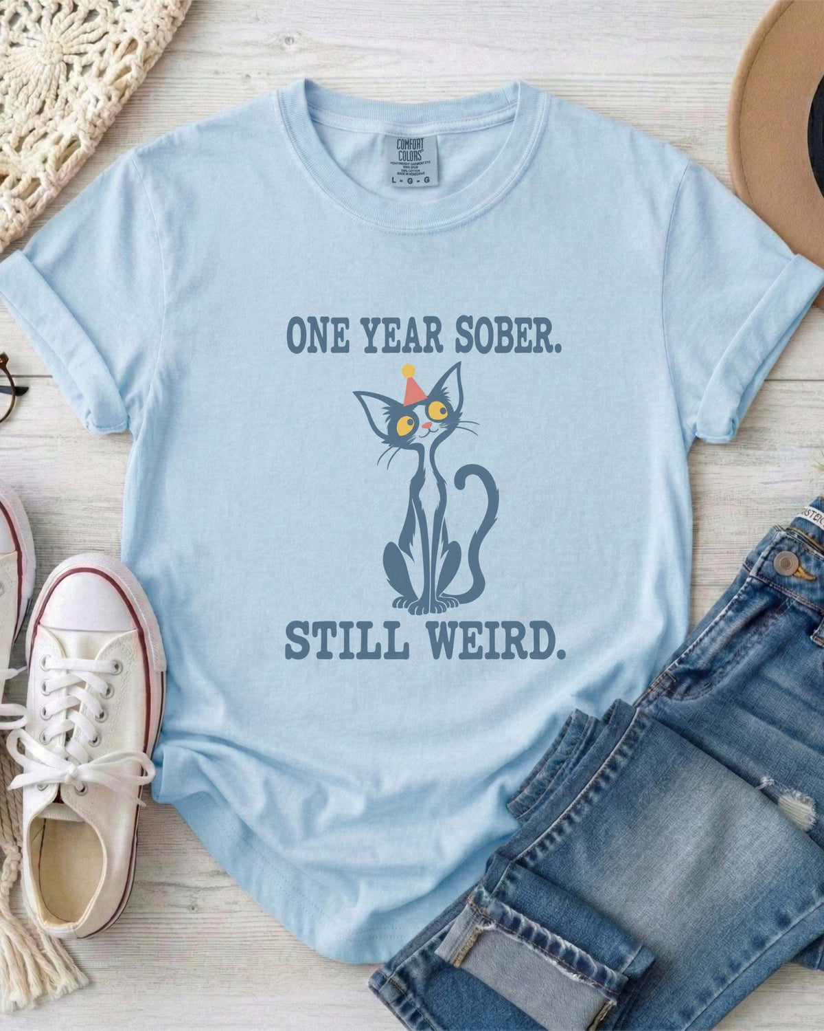 Flat lay of a chambray blue Comfort Colors 1717 t-shirt featuring a navy cat in a red party hat and “One Year Sober. Still Weird.” lettering.