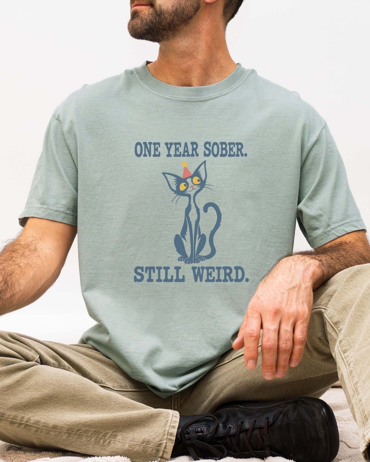 Male model wearing a bay green Comfort Colors 1717 t-shirt featuring a navy cat in a red party hat and “One Year Sober. Still Weird.” lettering.