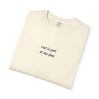 Folded beige t-shirt with text 'rest is part of the plan' on a white background