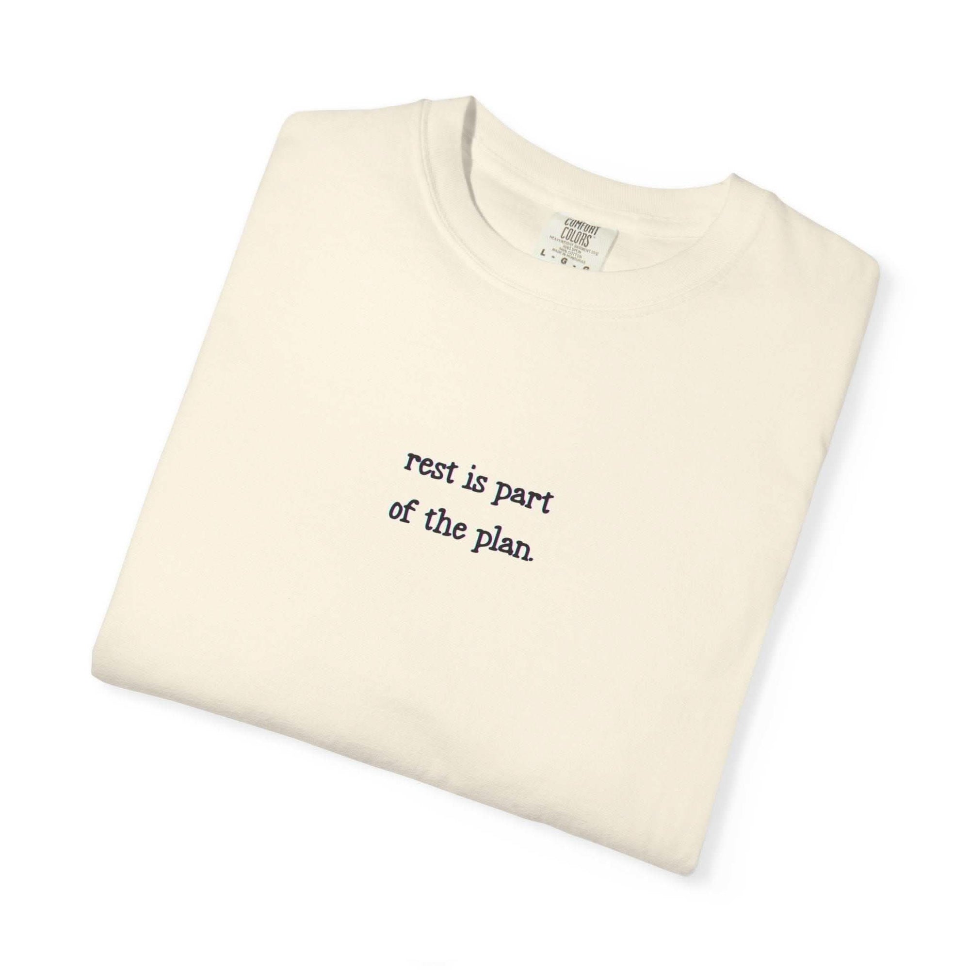 Folded beige t-shirt with text 'rest is part of the plan' on a white background