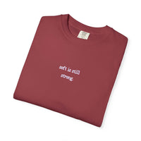 Folded maroon t-shirt with 'soft is still strong' text on a white background