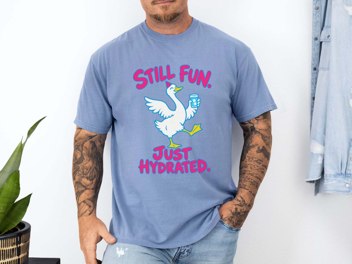 Male model wearing blue jean Comfort Colors crewneck t-shirt featuring a graphic of a white goose holding a blue water bottle, with the pink text 'STILL FUN. JUST HYDRATED.