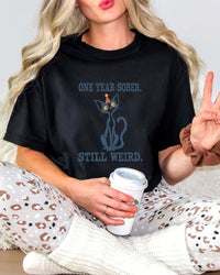 Female model wearing a black Comfort Colors 1717 t-shirt featuring a navy cat in a red party hat and “One Year Sober. Still Weird.” lettering.