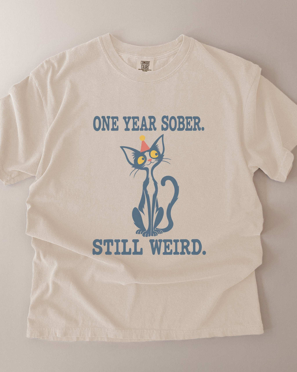 Flat lay mockup of an Ivory Comfort Colors 1717 t-shirt featuring a navy cat in a red party hat and “One Year Sober. Still Weird.” lettering.
