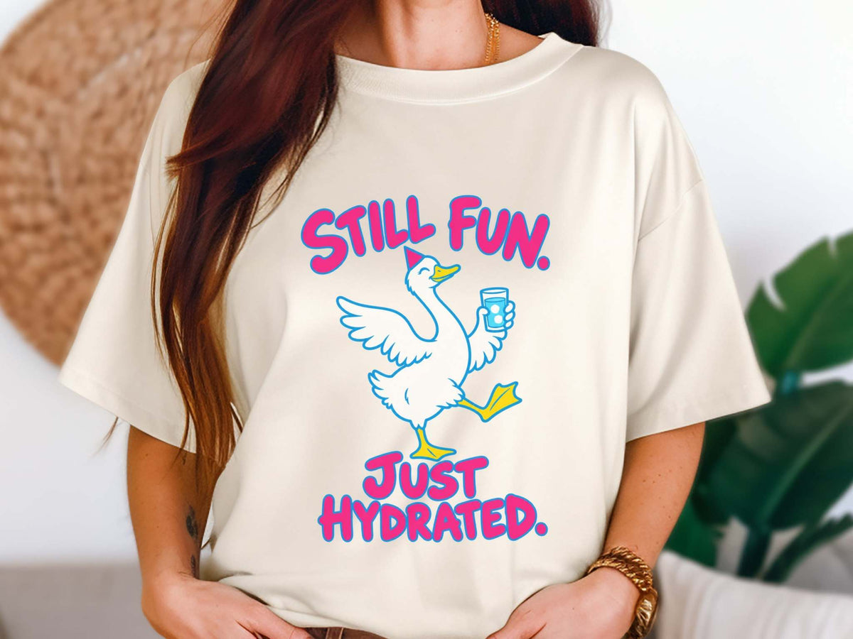 Female model wearing an ivory Comfort Colors crewneck t-shirt featuring a graphic of a white goose holding a blue water bottle, with the pink text 'STILL FUN. JUST HYDRATED.