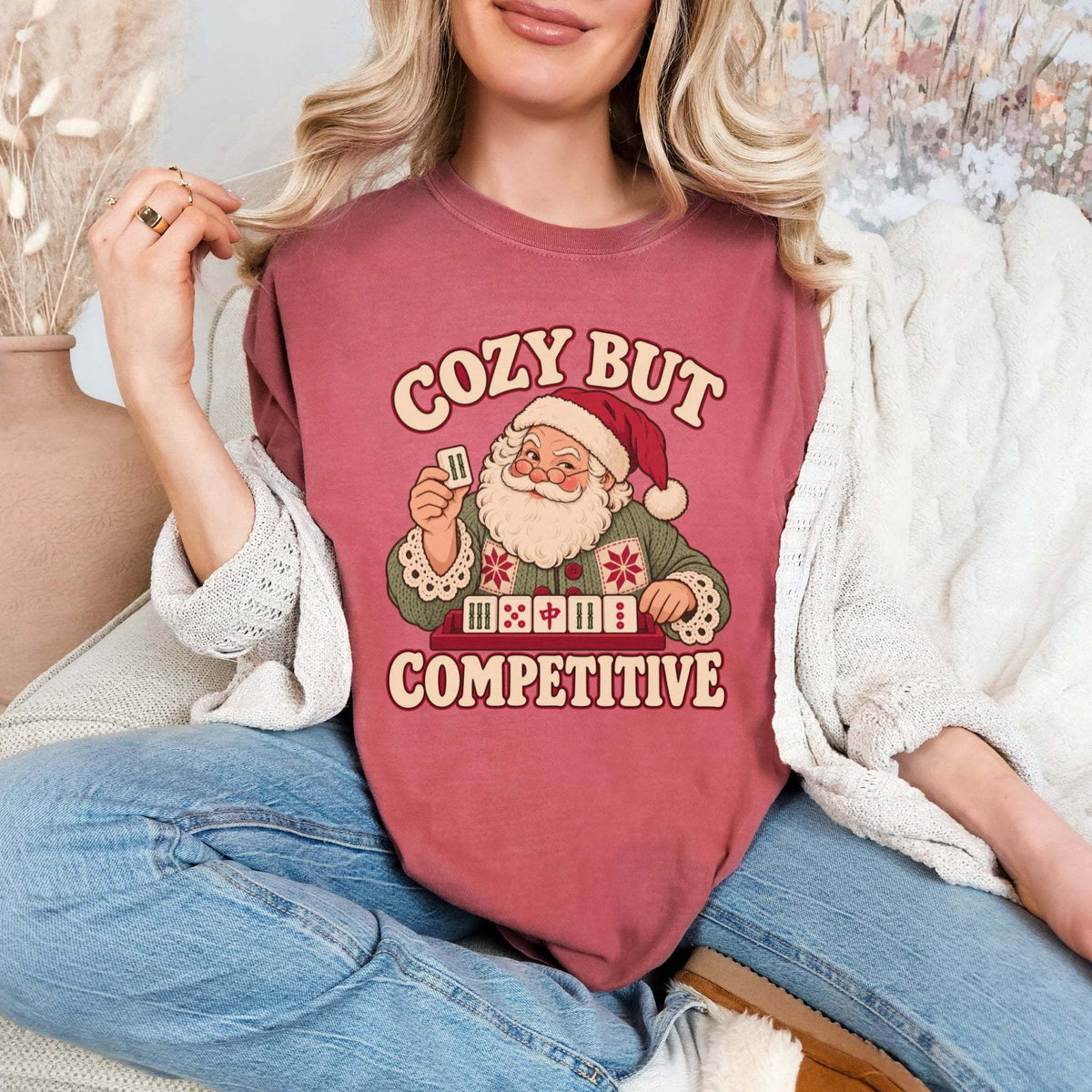Model wearing a Crimson red Comfort Colors T-shirt with Santa holding a mahjong tile above a tile rack and cream “Cozy But Competitive” lettering.
