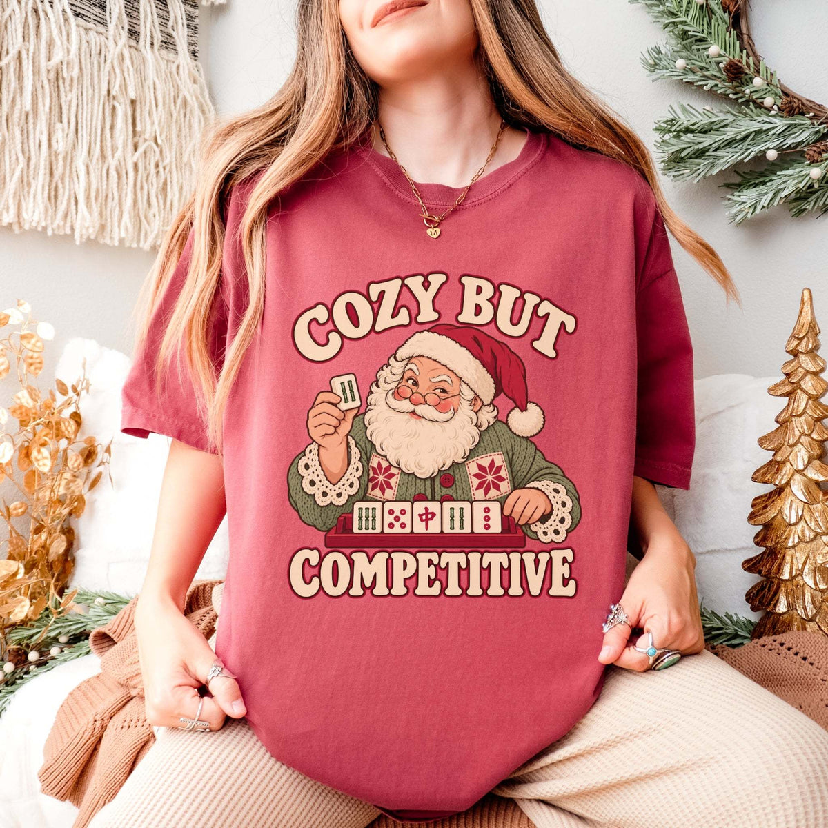 Model wearing a Crimson red Comfort Colors T-shirt with Santa holding a mahjong tile above a tile rack and cream “Cozy But Competitive” lettering.