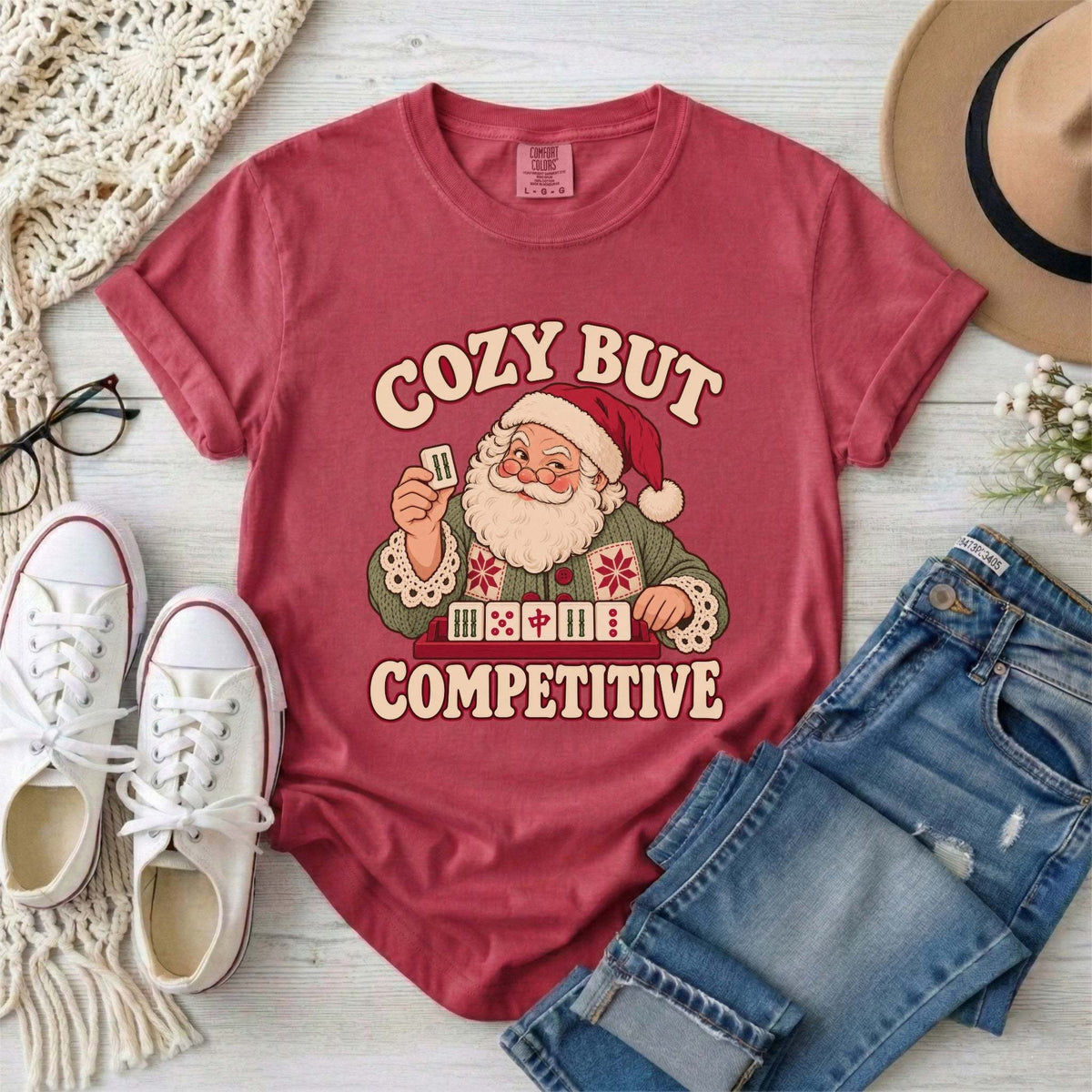 Flat lay of a Crimson red Comfort Colors T-shirt with Santa holding a mahjong tile above a tile rack and cream “Cozy But Competitive” lettering.