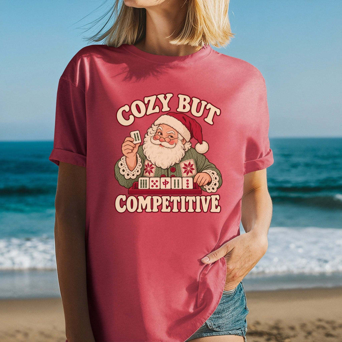 Model wearing a Crimson red Comfort Colors T-shirt with Santa holding a mahjong tile above a tile rack and cream “Cozy But Competitive” lettering.