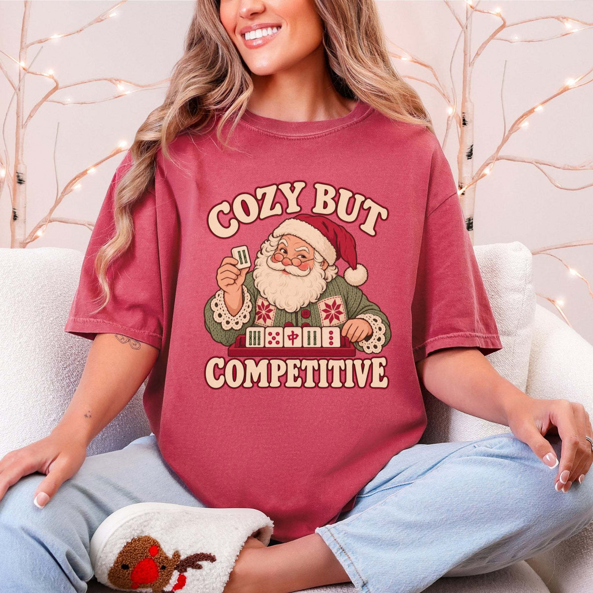 Model wearing a Crimson red Comfort Colors T-shirt with Santa holding a mahjong tile above a tile rack and cream “Cozy But Competitive” lettering.
