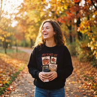 Woman holding coffee on a leaf-covered park path, wearing a black My Kind of Fall Plans graphic sweatshirt with blue jeans.