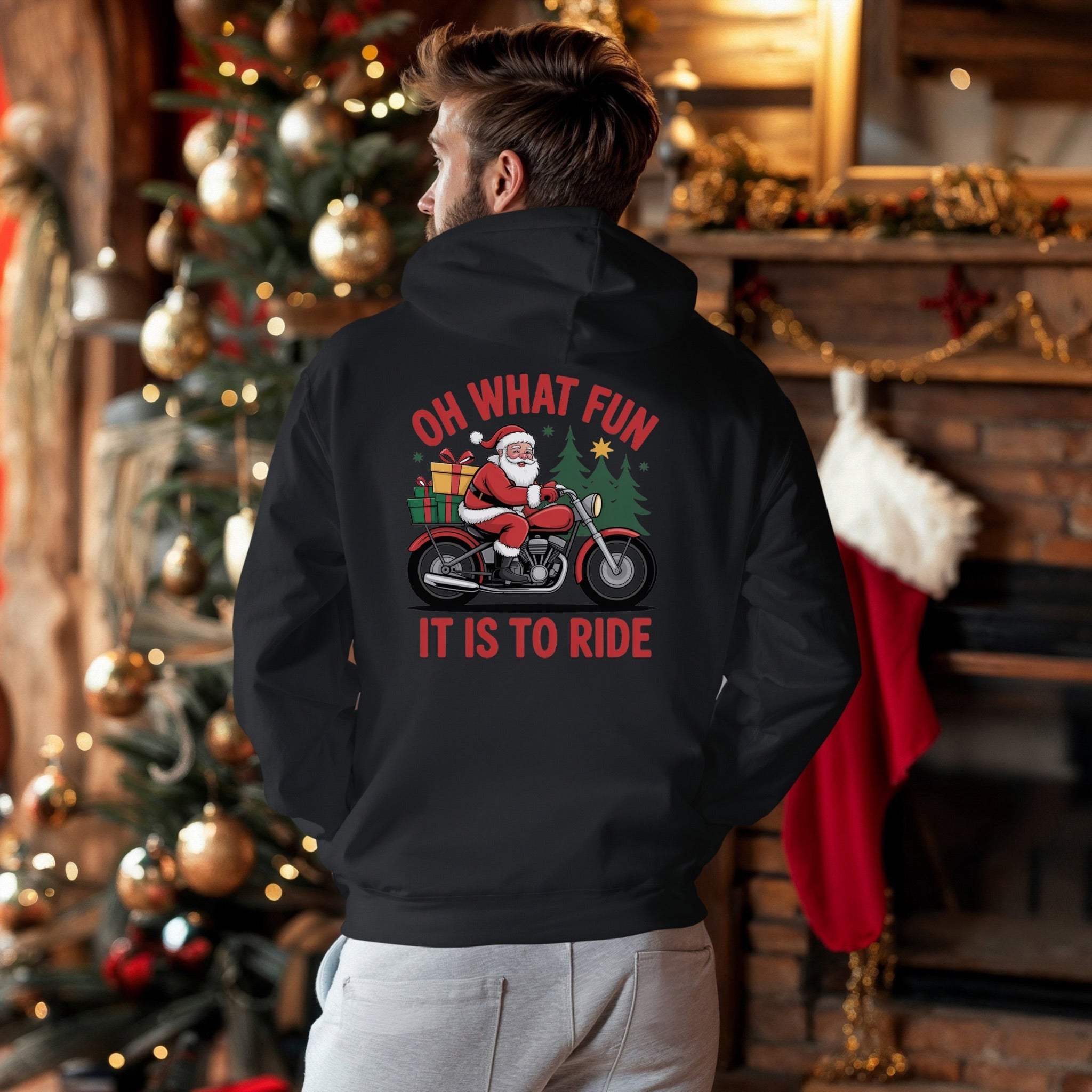 Man wearing black Santa Motorcycle Hoodie with Santa riding motorcycle and gifts graphic, festive holiday background with Christmas tree and stockings