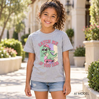 Girl wearing a grey skateboarding dinosaur T-shirt with “Catch Me If You Can” lettering and denim shorts on a sunny garden walkway.