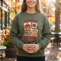 Woman wearing a military green My Kind of Fall Plans sweatshirt with coffee, maple leaf, book and cinnamon roll graphics outside an autumn café.