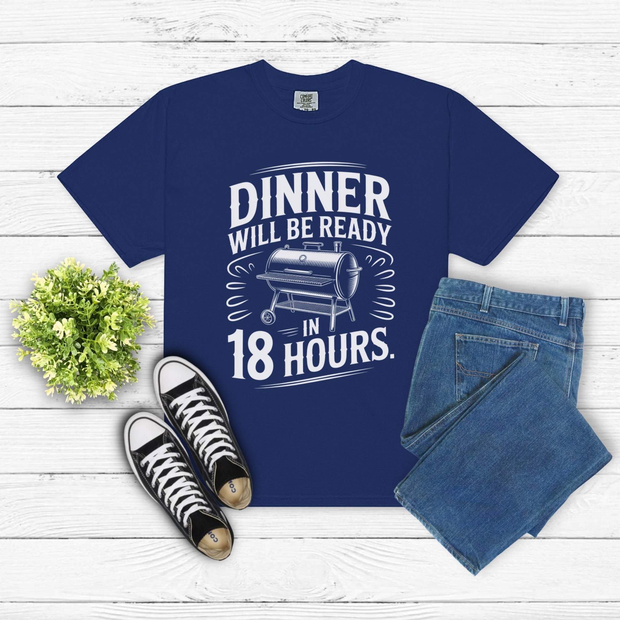 Dinner Will Be Ready in 18 Hours meat smoker t-shirt in navy with white text, paired with jeans and sneakers on white wood background