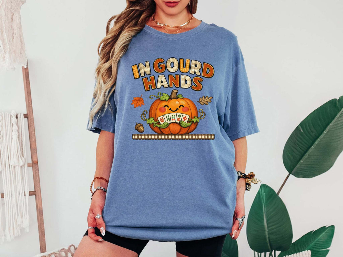 A blue jean short-sleeve t-shirt with a colorful graphic on the front. The graphic features the text 'IN GOUD HANDS' in a stylized orange and brown font above a smiling orange pumpkin with a toothy grin and autumn-themed details.