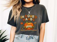 Model wearing a pepper short-sleeve t-shirt with a colorful graphic on the front. The graphic features the text 'IN GOUD HANDS' in a stylized orange and brown font above a smiling orange pumpkin with a toothy grin and autumn-themed details.
