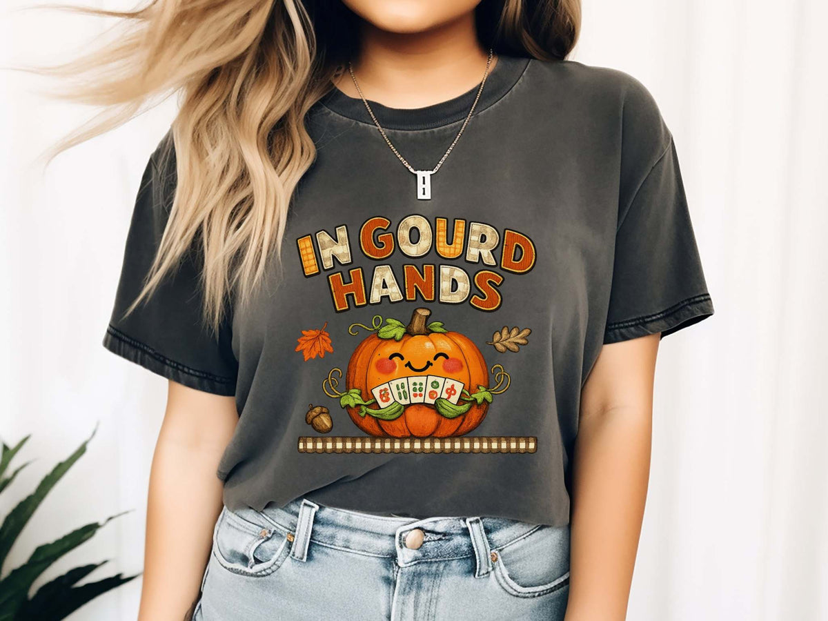Model wearing a pepper short-sleeve t-shirt with a colorful graphic on the front. The graphic features the text 'IN GOUD HANDS' in a stylized orange and brown font above a smiling orange pumpkin with a toothy grin and autumn-themed details.
